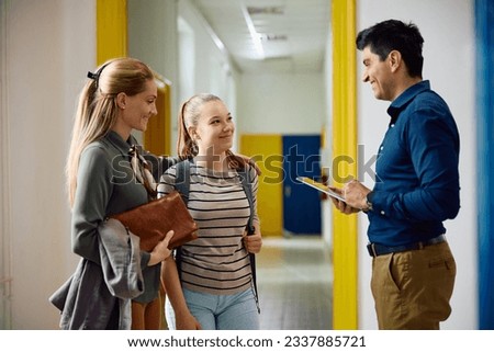 Happy mother and her teenage daughter communicating with a principal in high school hallway.