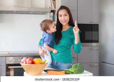 Happy Mother Holding Her Baby With One Finger Up Action On Kitchen Background. Mixed Race Asian-German Family Cooking Time With Many Vegetable On Table. Happy Mom And Kid.