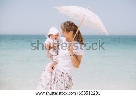 Happy mother holding her little baby girl on the beach in Dubai. In one hand she is holding a white parasol
