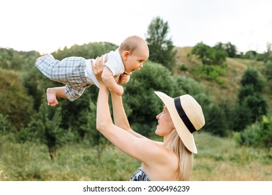 Happy Mother Holding Infant Baby Up In The Air Outdoors Wearing Sunhat At Summer Day