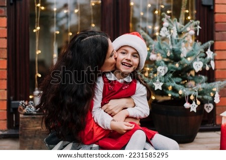 Happy mother hugging and kissing her laughing daughter while sitting on the porch of a house with decorated potted Christmas tree. Xmas mood. Family time. Close family relationship. Selective focus.