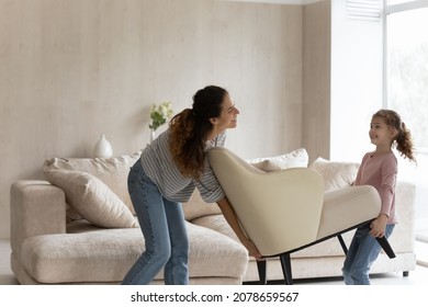 Happy mother with little daughter carrying armchair, smiling young mom and cute girl child decorating cozy living room in new own home apartment, moving day and relocation, renovation concept