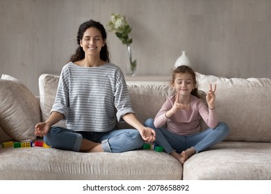 Happy Mother And Little Daughter Practicing Yoga, Meditating On Comfortable Couch At Home Together, Smiling Calm Mom Teaching Cute Girl Kid To Relaxing, Doing Exercises, Family Meditation Concept