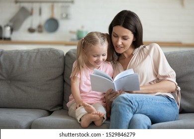 Happy Mother And Little Daughter Reading Book, Sitting On Comfortable Couch In Living Room, Smiling Mum Teaching Little Girl Child, Family Spending Weekend At Home Together, Children Education