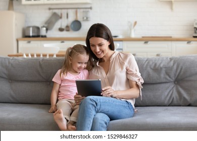 Happy mother and little daughter using computer tablet, making video call together, sitting on couch at home, smiling mum and preschool child chatting online, playing game, watching video