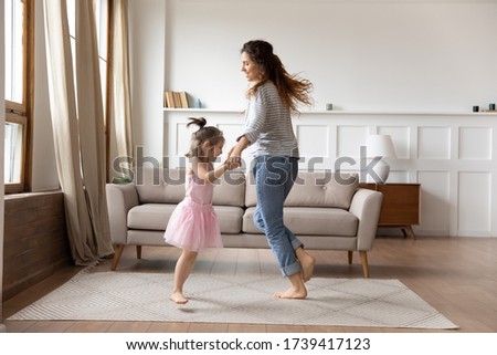 Happy mother and little daughter wearing princess dress holding hands, dancing in modern living room at home, family funny activity, smiling young mum and pretty preschool girl moving to music