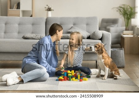 Happy mother and little kid playing with dog and colorful cubes at home, sitting on heating floor, stroking beloved pet, talking, laughing, smiling, building toy towers, castle