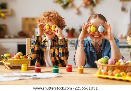 Happy mother and little son   holding painted colorful Easter eggs in front of eyes while decorating them with food dyes in cozy kitchen at home. Easter craft activities for families