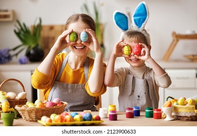 Happy mother and little son wearing aprons holding painted colorful Easter eggs in front of eyes while decorating them with food dyes in cozy kitchen at home. Easter craft activities for families