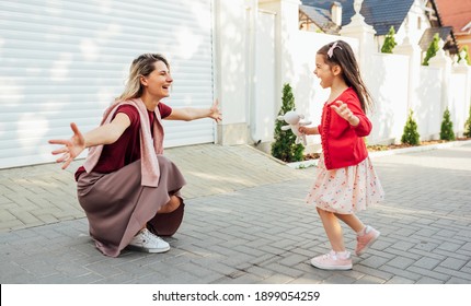 Happy Mother Meets Her Little Girl Going Back Home From School With Arms Open Outside. Happy Daughter Running To Her Smiling Mom Feeling Joyful After The Preschool Day. Happy Mother's Day.