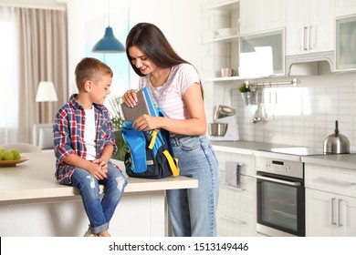 Happy Mother Putting Textbooks Into Little Child's School Bag In Kitchen