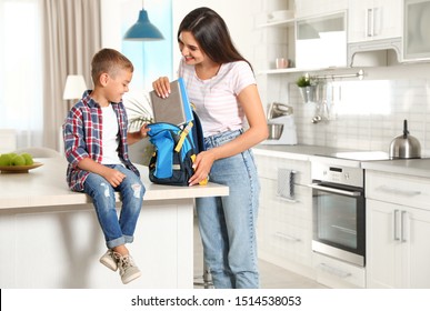 Happy Mother Putting Textbooks Into Little Child's School Bag In Kitchen