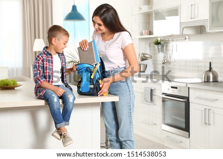 Happy mother putting textbooks into little child's school bag in kitchen