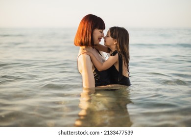 Happy Mother With Red Hair Holding Daughter In A Black Swimming Suit In The Sea. Happy Beautiful Mother And Daughter In Swimsuits Swimming In Sea, Family Look Concept.