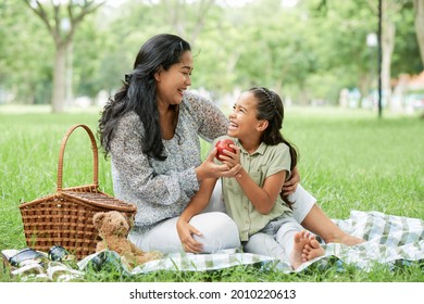 Happy mother sitting on the grass together with her little daughter and eating fruit during picnic in the park - Powered by Shutterstock