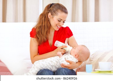 Happy Mother Sitting On Sofa And Feeding Baby