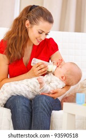 Happy Mother Sitting On Sofa And Feeding Baby