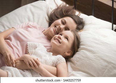 Happy mother with smiling cute little daughter relaxing in comfortable bed together, having fun, loving mum hugging cuddling adorable preschool child, enjoying morning in bedroom at home