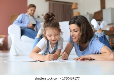Happy mother smilling daughter lying on warm floor enjoying creative activity, drawing pencils coloring pictures in albums, father resting on couch, family spend free time together.