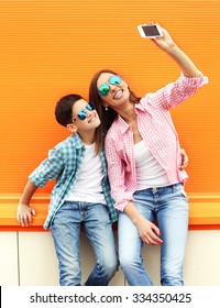 Happy Mother And Son Teenager Taking Picture Self Portrait On Smartphone In City, Over Colorful Background, Wearing A Checkered Shirt And Sunglasses 