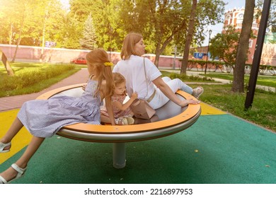 Happy Mother Spinning On Carousel With Two Girls On Modern Merry Go Round On Playground At Park