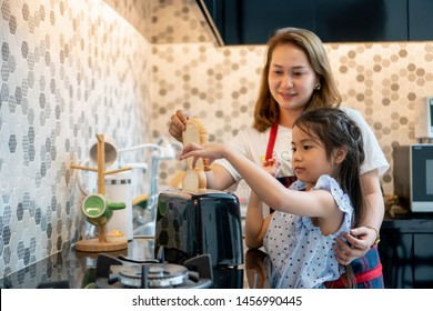 Happy Mother Teaching Daughter Making Breakfast Toast Bread With Toaster At Home Kitchen Together . Loving Family.  Child Girl Excited Looking Mom Cooking Indoors