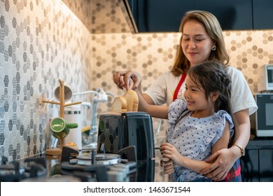 Happy Mother Teaching Daughter Making Breakfast Toast Bread With Toaster At Home Kitchen Together . Loving Family.  Child Girl Excited Looking Mom Cooking Indoors