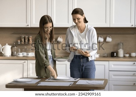 Happy mother and teen daughter child decorating table for dinner, placing dich, plates, talking, chatting, smiling, laughing, enjoying household activities. Kid helping mom with domestic chores