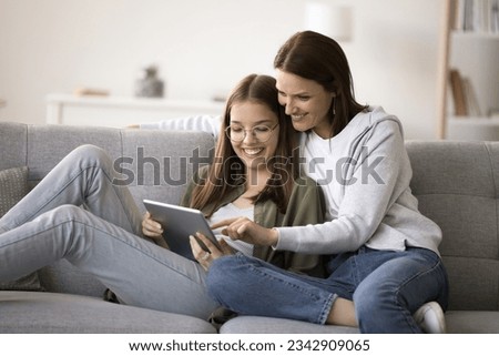 Happy mother and teenager kid girl using online application for shopping together, resting on sofa, holding digital tablet computer, smiling, laughing, enjoying domestic Internet communication