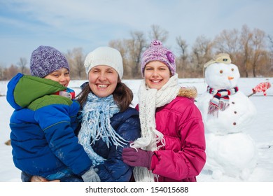 Happy Mother With Two Kids Building Snowman Outside In Winter Time