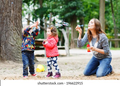 Happy mother and two little children playing together with soap bubbles on playground in summer
