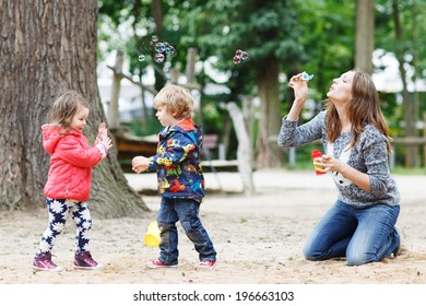 Happy mother and two little children playing together with soap bubbles on playground in summer