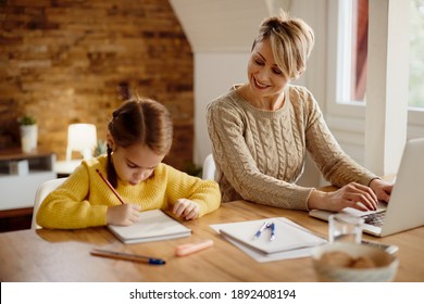 Happy mother using computer and working at home while daughter is writing homework beside her. 