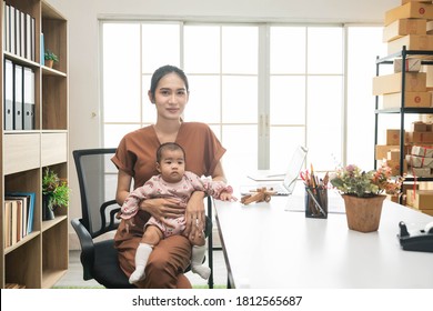 Happy Mother Working Online Holding Her Baby Daughter At Home Office. Single Mom Looking Camera With Her Child. Asian Woman Working From Home, While In Quarantine Isolation During Covid-19 Crisis.