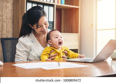 Happy Mother Working Online With A Laptop Holding Her Baby Son At Home Office. Smiling Single Mom With Her Child. Asian Woman Working From Home, While In Quarantine Isolation During Covid-19 Crisis.