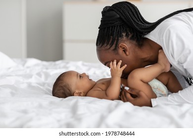 Happy Motherhood Concept. Side View Profile Portrait Of Cheerful Young Black Woman Playing With Her Adorable Small Baby Who Lying In Bed On White Bedsheets, Kissing Sweet Infant Child In Tummy