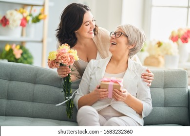 Gelukkige moederdag! Mooie jonge vrouw en haar moeder met bloemen en geschenkdoos thuis.                                