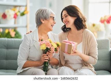 Happy Mother's Day! Beautiful Young Woman And Her Mother With Flowers And Gift Box At Home.                                