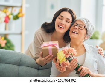 Happy Mother's Day! Beautiful Young Woman And Her Mother With Flowers And Gift Box At Home. 