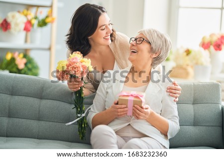 Happy mother's day! Beautiful young woman and her mother with flowers and gift box at home.                                