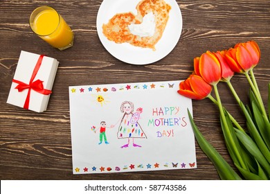 Happy Mothers Day Card Made By A Child. Mother's Hands Hold A Card A Gift From The Child And A Coffee Cup On A Wooden Background Of A Table.