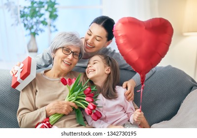 Happy Mother's Day! Child Daughter Is Congratulating Mom And Granny Giving Them Flowers Tulips. Grandma, Mum And Girl Smiling And Hugging. Family Holiday And Togetherness.