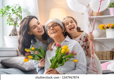 Happy Mother's Day! Child Daughter Is Congratulating Mom And Granny Giving Them Flowers Tulips. Grandma, Mum And Girl Smiling And Hugging. Family Holiday And Togetherness.