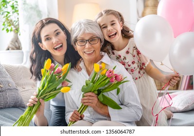 Happy Mother's Day! Child Daughter Is Congratulating Mom And Granny Giving Them Flowers Tulips. Grandma, Mum And Girl Smiling And Hugging. Family Holiday And Togetherness.