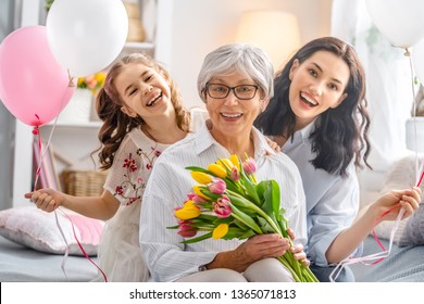 Happy Mother's Day! Child Daughter Is Congratulating Mom And Granny Giving Them Flowers Tulips. Grandma, Mum And Girl Smiling And Hugging. Family Holiday And Togetherness.