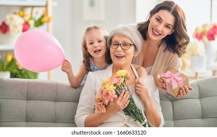 Happy Mother's Day! Child Daughter Is Congratulating Mom And Granny Giving Them Flowers And Gift. Grandma, Mum And Girl Smiling And Hugging. Family Holiday And Togetherness.