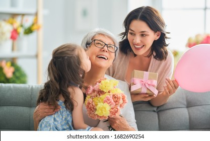 Happy Mother's Day! Child Daughter Is Congratulating Mom And Granny Giving Them Flowers And Gift. Grandma, Mum And Girl Smiling And Hugging. Family Holiday And Togetherness.                        