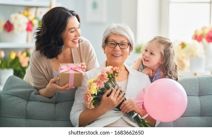 Happy Mother's Day! Child Daughter Is Congratulating Mom And Granny Giving Them Flowers And Gift. Grandma, Mum And Girl Smiling And Hugging. Family Holiday And Togetherness.