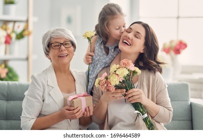 Happy Mother's Day! Child Daughter Is Congratulating Mom And Granny Giving Them Flowers And Gift. Grandma, Mum And Girl Smiling And Hugging. Family Holiday And Togetherness. 