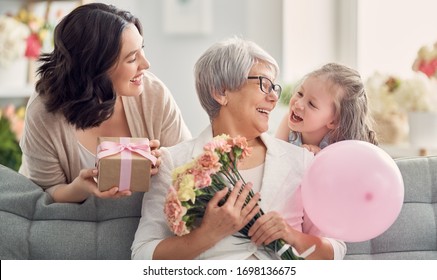 Happy Mother's Day! Child Daughter Is Congratulating Mom And Granny Giving Them Flowers And Gift. Grandma, Mum And Girl Smiling And Hugging. Family Holiday And Togetherness. 